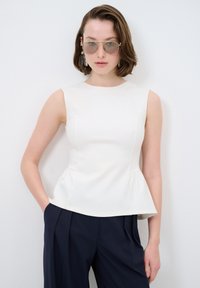 Weißes, tailliertes ärmelloses Oberteil mit einem Peplum-Saum, aus einem glatten Stoff, kombiniert mit marineblauen Hosen. Das Model steht mit den Händen in den Taschen.