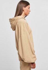 Beige színű, oversized kapucnis pulóver, húzószalagos kapucnival, hosszú ujjakkal és laza szabással. Puha anyag, sima tapintással.
