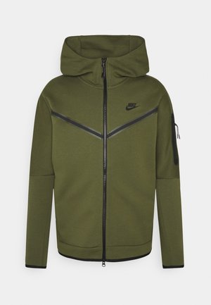 Oliivinvihreä Nike vetoketjullinen huppari, jossa on huppu, musta sahalaitakuosi rinnassa, Nike-logo vasemmassa rinnassa ja vetoketjullinen tasku vasemmassa hihassa.