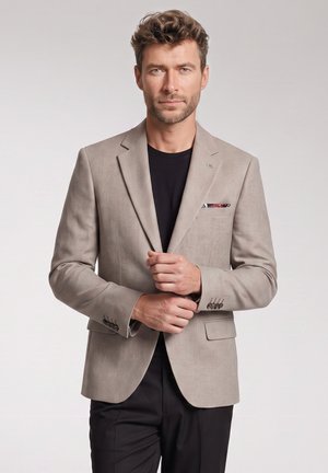 Man with short curly hair wearing a beige blazer over black shirt and black pants, adjusting the blazer buttons, standing against plain background.