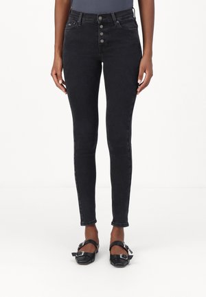 Femme portant un jean skinny noir taille haute avec une braguette à boutons et des ballerines noires à bride boucle, debout sur fond blanc.