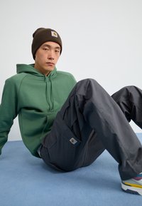 Hoodie vert, pantalon cargo gris. Tissu doux, coupe décontractée, logo visible sur le pantalon. Personne assise sur fond bleu, portant un bonnet marron.