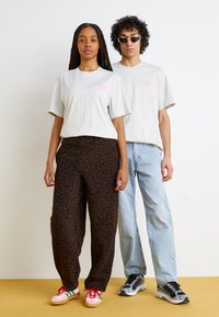 Ljusgrå t-shirts med rosa logotyp, bruna leopardmönstrade byxor och olika sneakers. Bakgrunden är enkel och ljus.