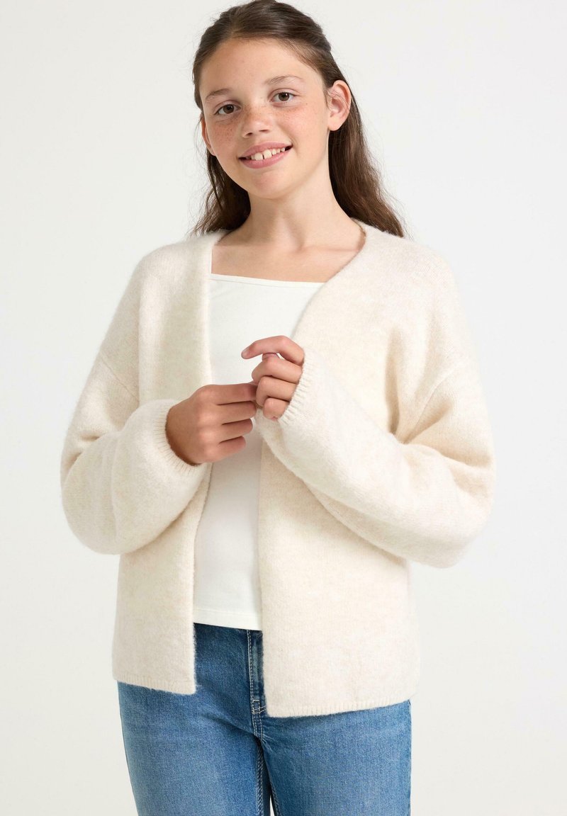 Lys beige cardigan lavet af blødt, fluffy materiale. Den har lange ærmer, en åben front og en afslappet pasform. Båret over en hvid top.
