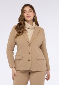 Donna che indossa un blazer beige con borchie e bottoni sopra un maglione a collo alto beige e pantaloni abbinati, in piedi contro uno sfondo semplice.