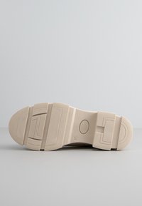 Sole in gomma beige con grip testurizzato, caratterizzata da più motivi rettangolari e un'emblema circolare, design piatto e largo.