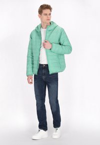 Lichtgroene pufferjack met een capuchon, ritsluiting en gewatteerde textuur, gecombineerd met donkerblauwe jeans en witte sneakers.