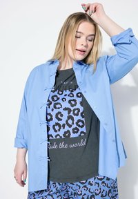 Helles blaues Hemd mit Rüschendetail auf der Vorderseite über einem schwarzen T-Shirt mit blauem Leopardenmuster und dem Text "Die Welt regieren."