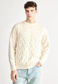 Krém színű, kötött mintájú pulóver kerek nyakkivágással és bordázott mandzsettákkal. Világoskék farmernadrág fölött viselve.