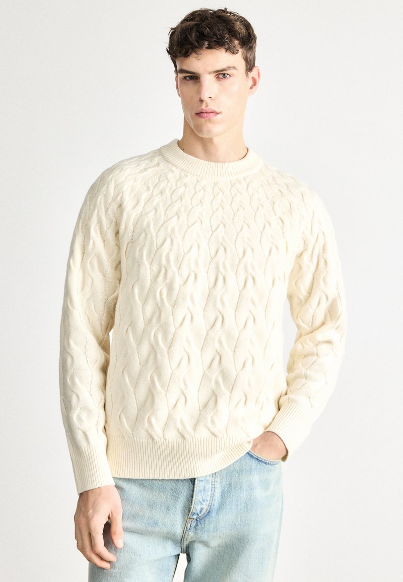 Krém színű, kötött mintájú pulóver kerek nyakkivágással és bordázott mandzsettákkal. Világoskék farmernadrág fölött viselve.