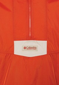 Gros plan sur le tissu orange d'une veste avec une fermeture éclair verticale et un patch rectangulaire blanc portant l'inscription Columbia Sportswear Company.