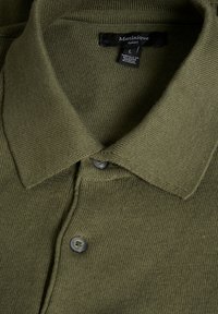 Polo de color verde oliva con un tejido texturizado, cierre de botones y diseño de cuello clásico. La etiqueta muestra la talla L y el nombre de la marca.