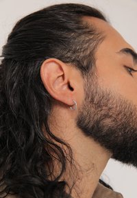 Silverfärgad creolörhänge, cirkulär form, slät metallfinish, bärs i ett piercat öra. Håret är långt, mörkt och vågigt, inramande ansiktet.