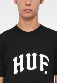 Fekete pamut póló rövid ujjakkal és kerek nyakkivágással. Az elején nagy, fehér grafikus "HUF" felirat található. Simótextúra.