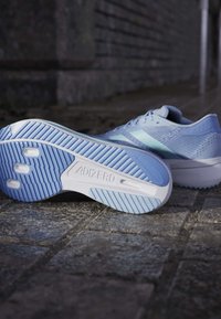 Helles blaues Sportschuh mit strukturiertem Obermaterial und weißer Sohle; verfügt über auffällige ADIZERO-Branding und ein gemustertes Profil für Traktion.