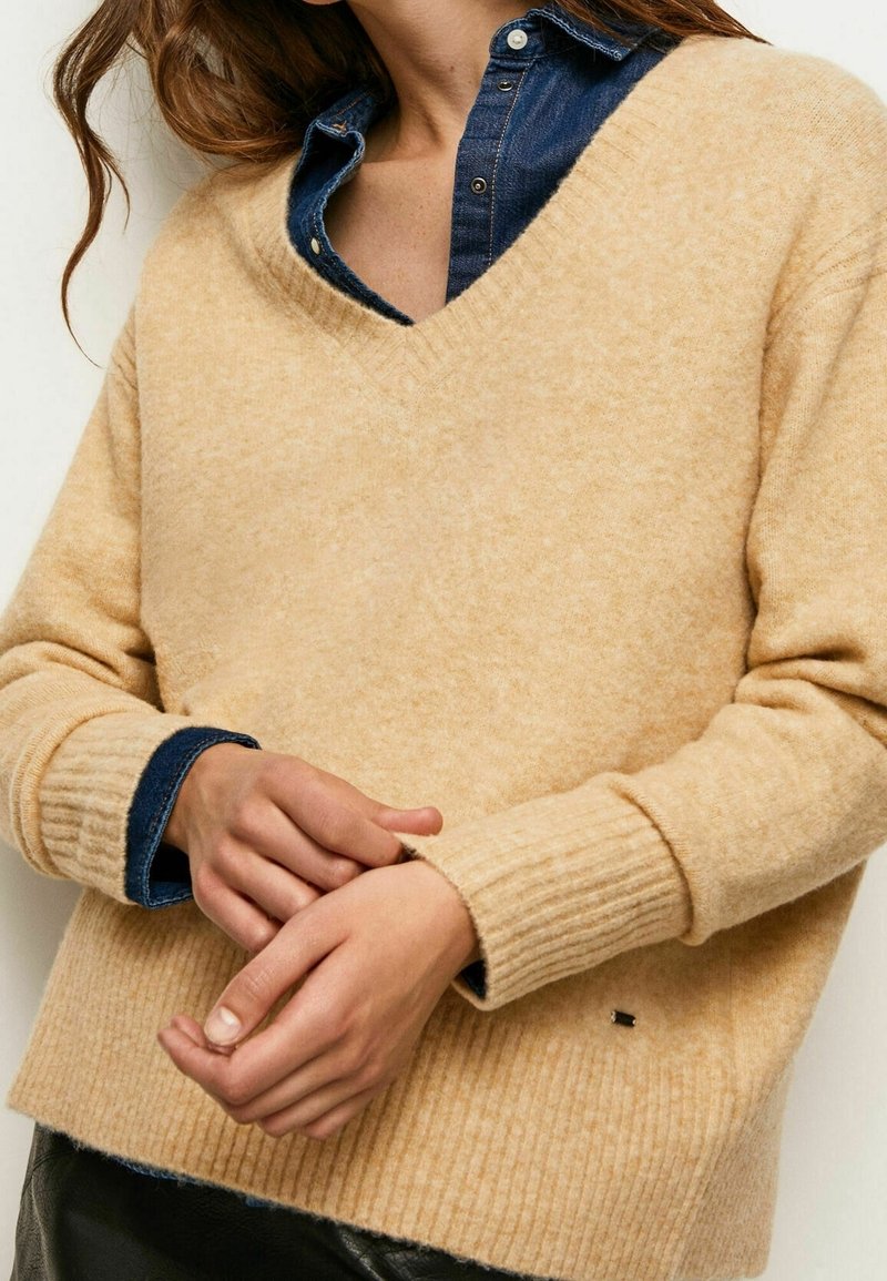 Pull à col en V beige sable fabriqué dans un matériau texturé avec des poignets côtelés. Porté sur une chemise en denim avec un petit détail de logo.