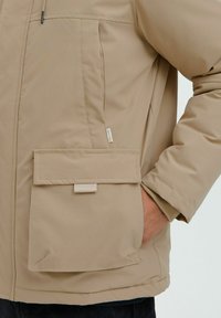Beige padded jacket featuring a large flap pocket, drawstring details at the collar, and contrasting fabric at the cuffs.