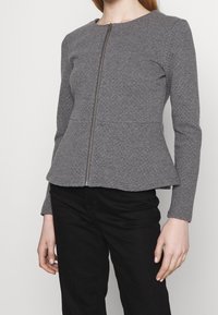 Grå peplumjacka med texturerad tyg, långa ärmar och frontdragkedja. Designad för en figursydd silhuett med rund halsringning.