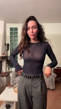 Schwarzes, transparentes Langarmoberteil kombiniert mit hoch taillierter, weit geschnittener grauer Hose. Schwarzer Gürtel mit metallischen Ösen als Akzent. Neutraler Hintergrund.