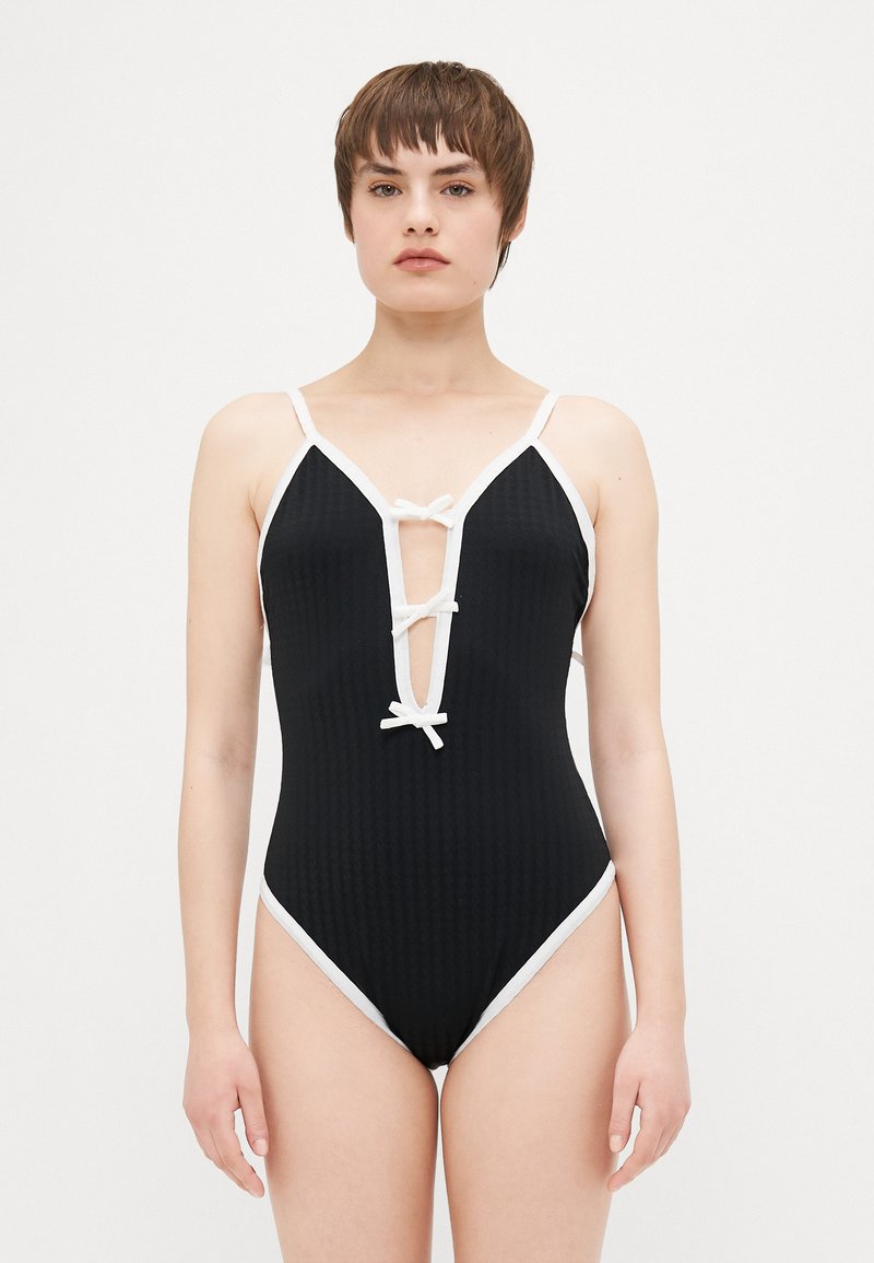 Maillot de bain une pièce noir avec une texture à côtes, accents blancs, décolleté en V profond et liens décoratifs à l'avant. Bretelles réglables.