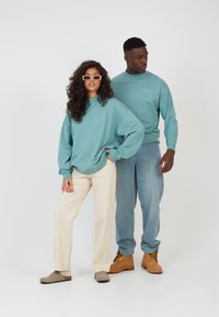 To personer iført matchende teal hettegensere med logo-detaljer. Kvinnen kombinerer sin med beige bukser og slip-on sko; mannen har lyseblå jeans og støvler.