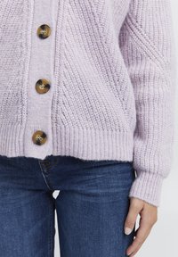 Cardigan lavorato a maglia di un leggero colore viola con un motivo testurizzato, dotato di cinque grandi bottoni marroni e polsini a coste, abbinato a jeans blu scuro.
