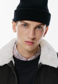 Berretto a coste nero, giacca di jeans con colletto in ecopelliccia bianca, indossato sopra un maglione nero, con una camicia a quadri che sbuca sotto.