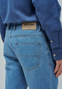 Jeans de mezclilla azul claro con bolsillos traseros y una etiqueta marrón que dice "SALSA JEANS", llevados con una camisa de manga larga azul oscuro.