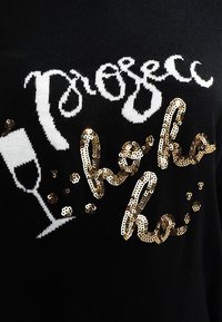 Schwarzer Strickpullover mit weißen und goldenen Paillettenverzierungen. Der Text lautet "Prosecco" mit einer Grafik eines Sektglases. Glitzernde Textur.