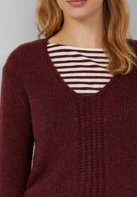 Eine Frau trägt einen burgunderfarbenen Strickpullover über einem weißen und burgunderfarben gestreiften Hemd, mit schulterlangem, hellbraunem Haar.