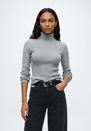 Kvinne med langt svart hår iført en grå ribbestrikket turtleneck-genser og svarte jeans med svart belte, stående foran en ensfarget bakgrunn.