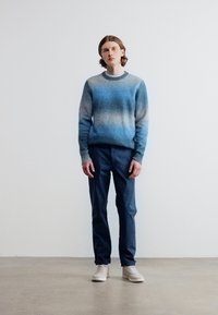 Νεαρό άτομο που στέκεται μπροστά σε απλό τοίχο, φορώντας μπλε πουλόβερ με διαβάθμιση, σκούρο μπλε παντελόνι και λευκά αθλητικά παπούτσια.