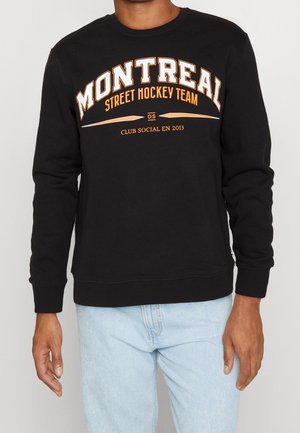 Mężczyzna w czarnej bluzie z napisem "Montreal Street Hockey Team Club Social en 2013", w jasnoniebieskich dżinsach, stoi na białym tle.