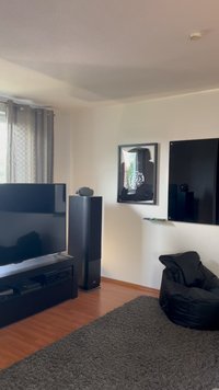 Móvel de TV preto com grande tela plana, altifalante adjacente, tapete felpudo cinza e cadeira de feijão preta; obra de arte emoldurada na parede branca.