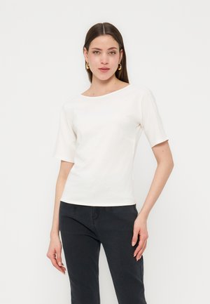 Femme portant un haut blanc à manches courtes et un pantalon noir, debout devant un fond clair uni, avec des boucles d'oreilles créoles dorées.