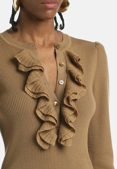 Brūns ribots kardigans ar viļņotu priekšpusi, kuram ir lielas zeltainas un baltas pogas. Audumam ir mīksta tekstūra, un tam ir pielāgota forma ar delikātu spīdumu.