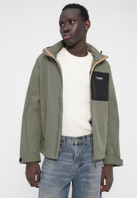 Giacca verde oliva con tasca nera a contrasto, chiusura con cerniera e dettagli marroni; abbinata a una maglietta bianca a coste e jeans blu.