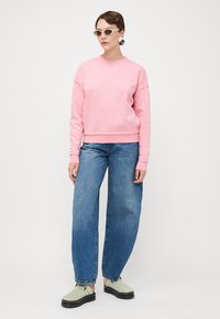 Felpa rosa con maniche borchiate, vestibilità rilassata, abbinata a jeans a gamba larga blu e scarpe slip-on verdi. Il modello indossa occhiali da sole.