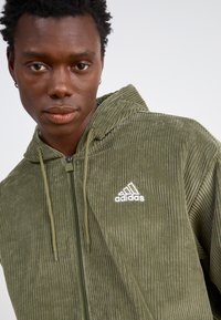 Olīvzaļš korduroja apvalks ar rāvējslēdzēju, kuram ir kapuce, ar baltu Adidas logotipu, teksturētu ribotu audumu un auklu.