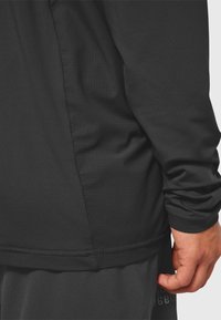 Prawa ręka i ramię osoby ubranej w czarny, długorękawowy strój z teksturowanymi panelami materiału oraz dopasowane ciemne spodnie.