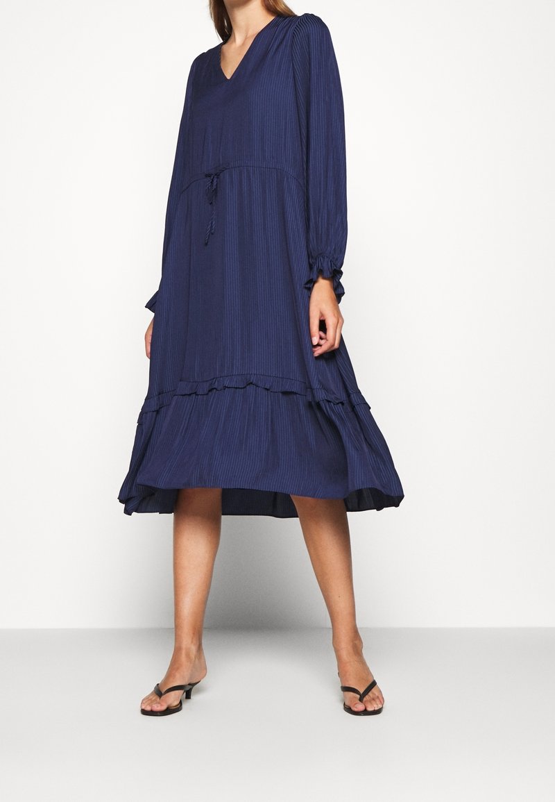 Námořnický modrý šaty ke kolenům s vertikálními sklady, výstřihem do V, dlouhými rukávy, stahovací pas a volánkovým lemem.