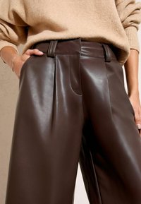 Moteris vilki tamsiai rudus odinius platėjančius kelnes su aukštu liemeniu ir šviesiai rudą purų megztinį, rankas ilsinanti arti kišenių.