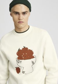 Pull crème avec un motif tricoté représentant un bol de fruits rouges. Encolure et poignets côtelés ; texture lisse, style ajusté.