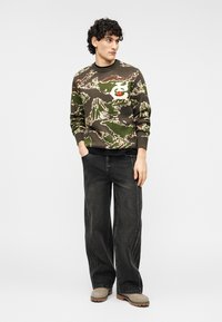 Πουλόβερ σε καμουφλαρισμένο μοτίβο, ελιάς, με γραφικό αρκούδας, συνδυασμένο με χαλαρά, σκούρα γκρι τζιν και μπεζ παπούτσια. Καθημερινό στυλ.