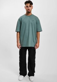 Grønn oversized t-skjorte med korte ermer, rund hals og subtil logo; kombinert med svarte cargo-bukser med flere lommer. Hvite sneakers.
