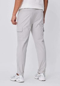 Homme portant un pantalon cargo gris clair avec des poches à rabat, des chaussures de sport blanches et une chemise blanche, debout devant un fond clair uni.