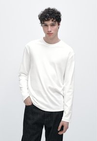 Weißes Langarm-T-Shirt mit rundem Ausschnitt, glattem Stoff und lockerem Schnitt, kombiniert mit dunklen Slim-Fit-Jeans.