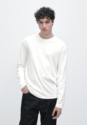 Hvid langærmet t-shirt med rund halsudskæring, glat stof og afslappet pasform, kombineret med mørke slim-fit jeans.