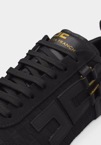 Μαύρο δερμάτινο αθλητικό παπούτσι με μαύρα κορδόνια και χρυσό λογότυπο "DC" και κείμενο στη γλώσσα, με γεωμετρικές λεπτομέρειες στο πλάι και χρυσές πινελιές.