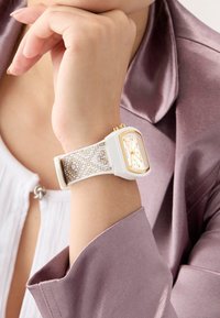Montre rectangulaire blanche avec des accents dorés et un bracelet texturé beige. Le cadran de la montre présente plusieurs cadrans et un affichage clair.